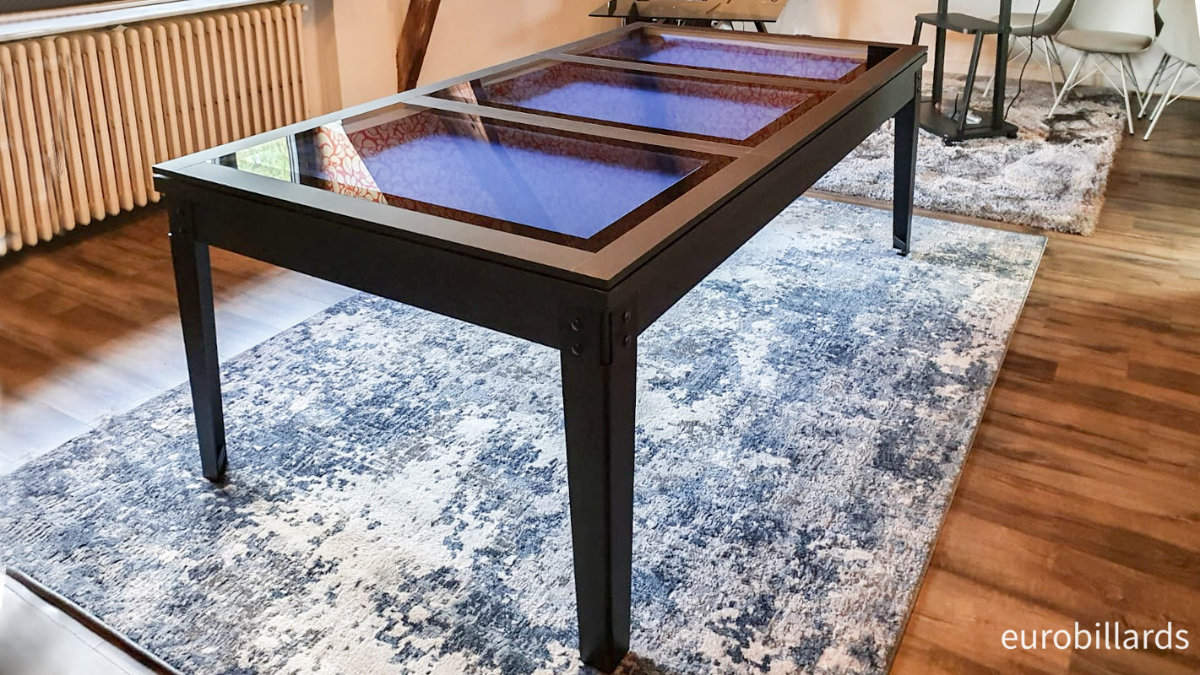 Billard transformable en table noire de style industriel avec plateau en verre qui laisse apparaitre le tissu de jeu américain couleur bleu roi, dont les lignes perpendiculaires formées par le cadre nano graphite, les pieds en métal noir et les bandeaux des verres renforcent avec élégance le côté ludique de cette salle de loisir.